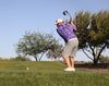 Golfer in a purple shirt and gray shorts swinging a golf club on a golf course with trees in the background.