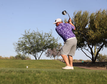 Golfer in a purple shirt and gray shorts swinging a golf club on a golf course with trees in the background.