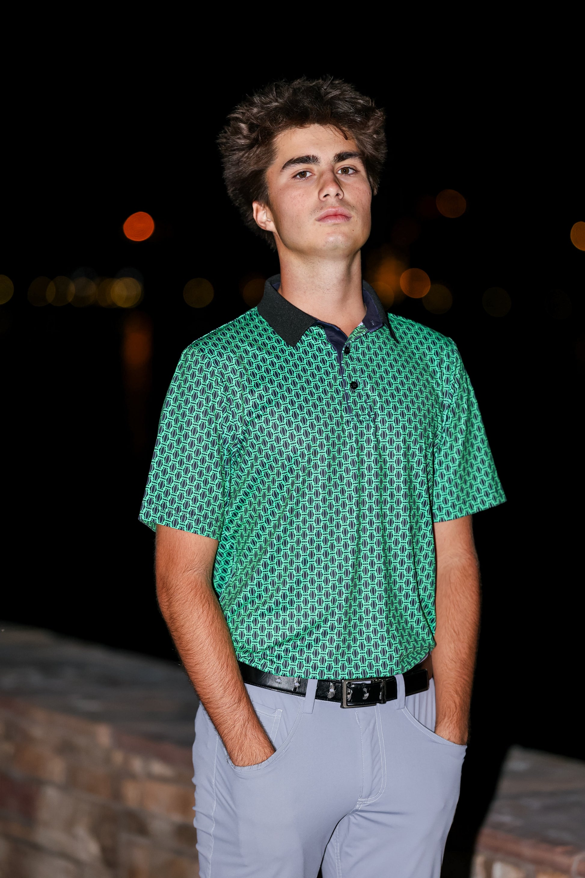 Man wearing a green patterned shirt and gray pants standing against a dark background